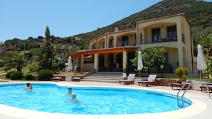 A swimming pool with lounge chairs and umbrellas, surrounded by a building with a balcony and a mountainous backdrop. - Exceptional villa: 400 m²  for 15 people, with pool (Tertenia)