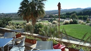 An outdoor seating area with tables and chairs, a palm tree, and a view of a green landscape. - First Floor Apartment On Golf Resort, Shared PoolWell Equipped Apartment (VERA)