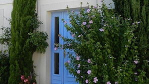 Een gebouw met twee verdiepingen, met een blauwe deur, een klein balkon en een tuin vol planten en bloemen.