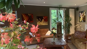 Een woonkamer met een bruine bank, een salontafel en een grote pot met roze bloemen. Twee schilderijen hangen aan de muur en er is een raam met uitzicht op groen.