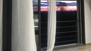 Une pièce dotée d’une grande porte-fenêtre coulissante, de rideaux blancs et offrant une vue sur un coucher de soleil se déclinant sur un paysage.