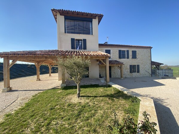 A two-story stone building with a tiled roof, a wooden pergola, and a small tree in the foreground.