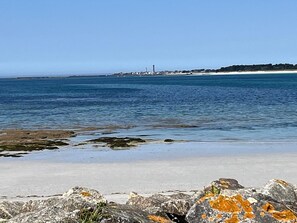 A coastal beach with a lighthouse in the distance, rocky foreground, and clear blue sky.
