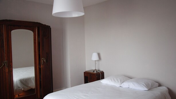 Ein Schlafzimmer mit einem Holfischschrank, einem Nachttisch mit Lampe und einem Bett mit weißer Bettwäsche.