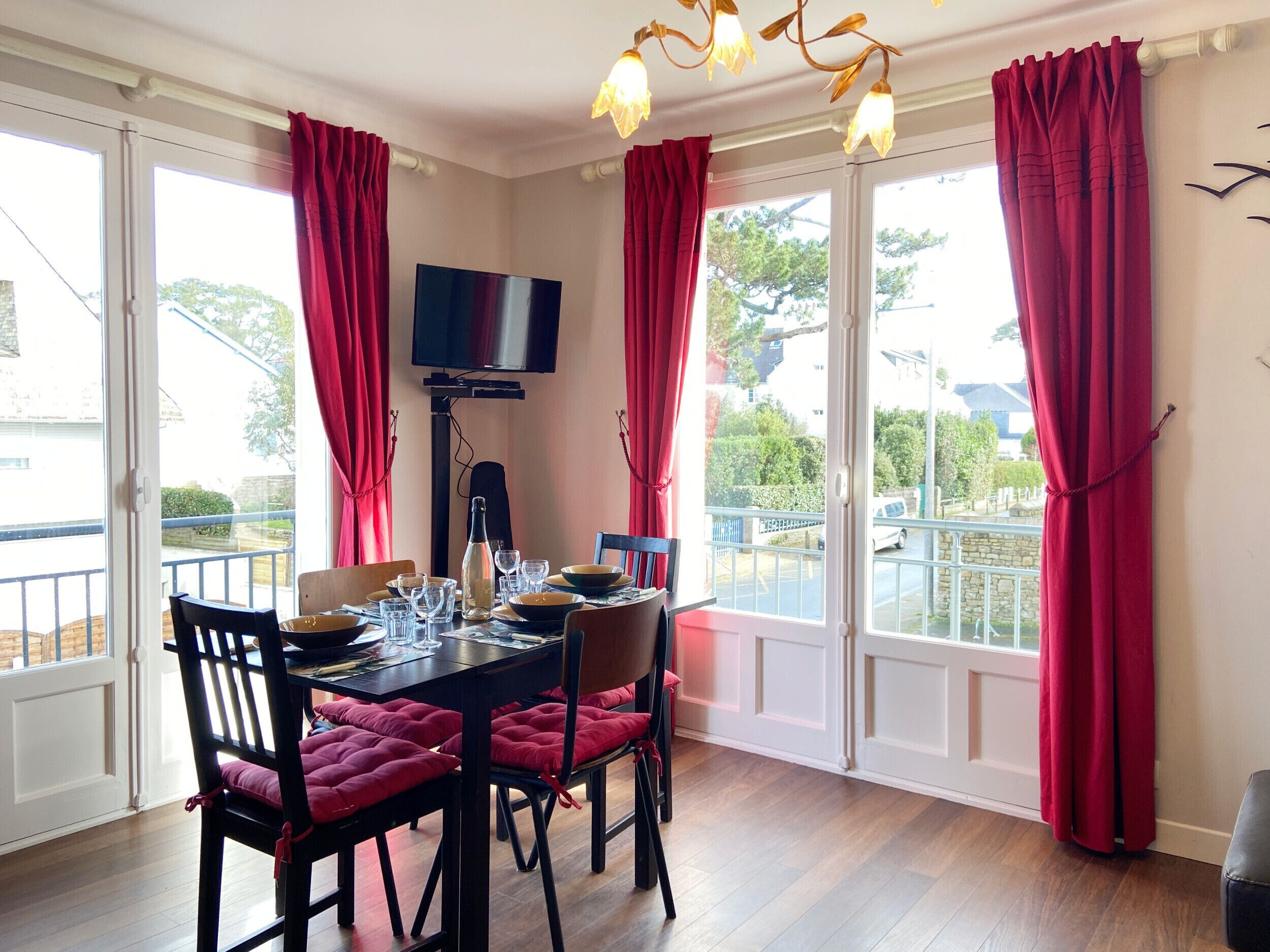 A dining area with a table set for four, a television mounted on the wall, and large windows with red curtains.