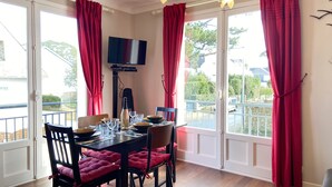 Un comedor con una mesa dispuesta para cuatro personas, un televisor montado en la pared y grandes ventanales con cortinas rojas.