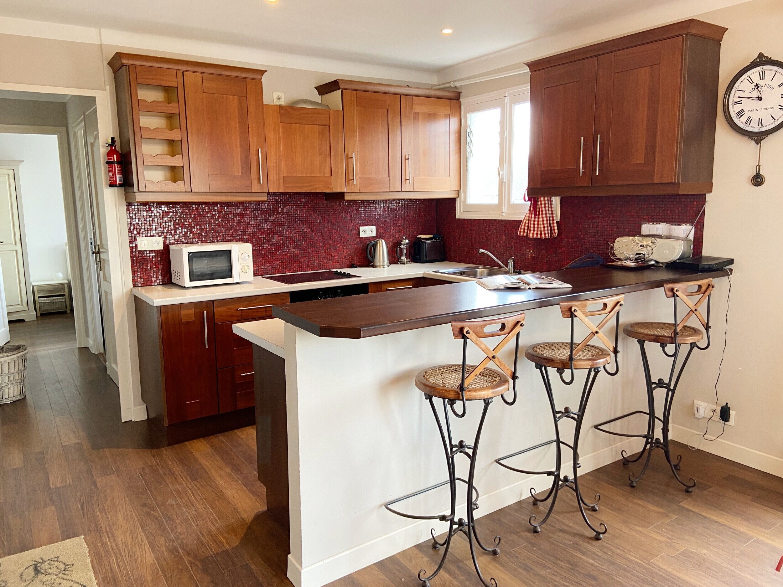 A kitchen with wooden cabinets, a white countertop, a microwave, a kettle, and a clock on the wall.