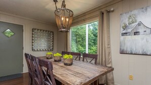 A dining room with a wooden table, chairs, a chandelier, and a window with a view of greenery.