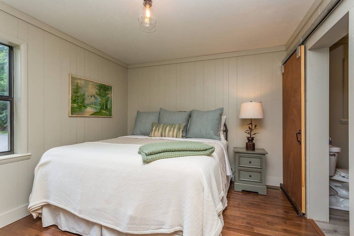 A bedroom with a bed, bedside table, lamp, painting, and a door leading to a bathroom.