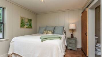 A bedroom with a bed, bedside table, lamp, painting, and a door leading to a bathroom.