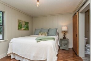 Une chambre à coucher comprenant un lit, une table de chevet, une lampe, un tableau et une porte donnant sur une salle de bain.