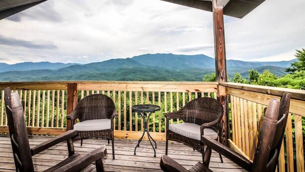 A wooden deck with wicker chairs and a small table, overlooking a mountainous landscape.