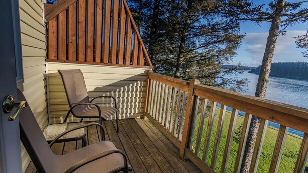 Une terrasse en bois avec deux chaises et une petite table, donnant sur un lac bordé d’arbres et sous un ciel dégagé.