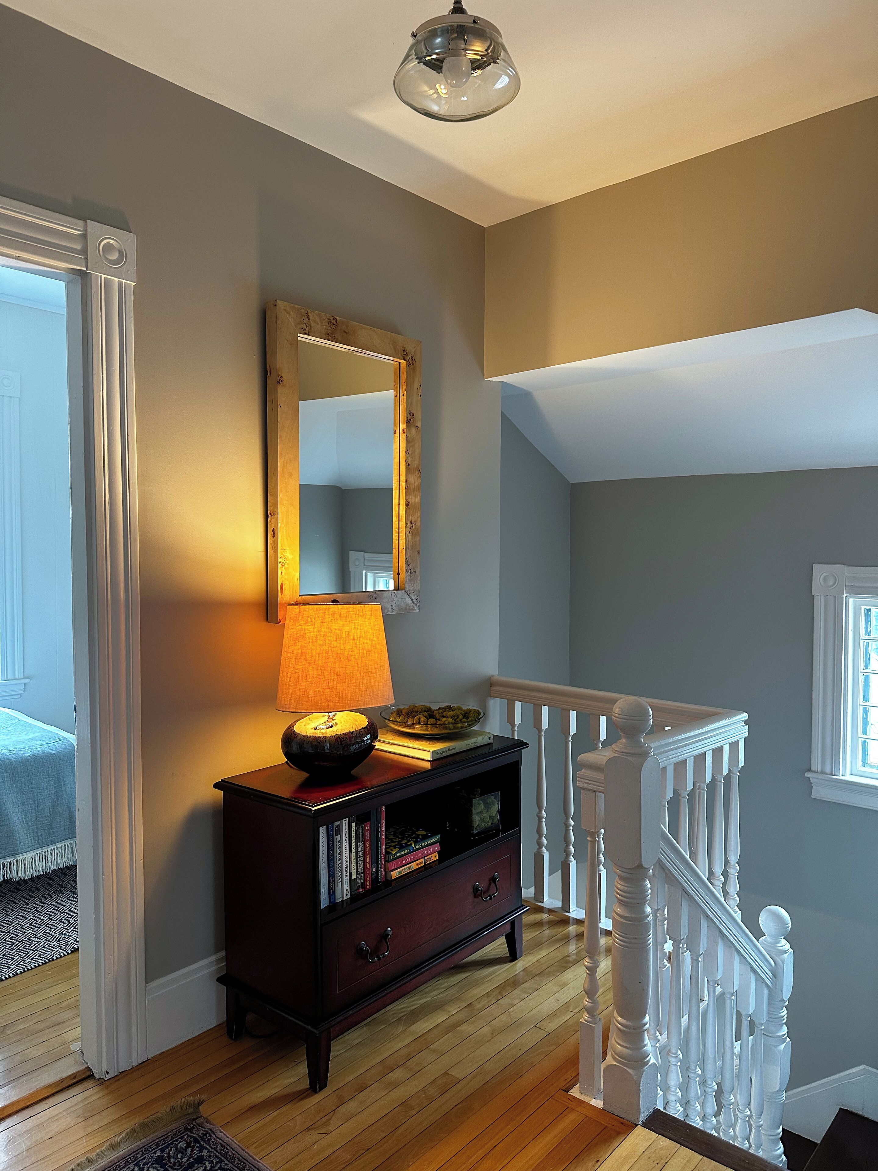 A wooden floor, a dark wooden console table with a lamp, a mirror, and a staircase with white balusters.