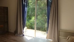 Une pièce dotée d’un plancher en bois, d’une table blanche, d’un canapé et d’une porte-fenêtre coulissante donnant sur un espace extérieur verdoyant.