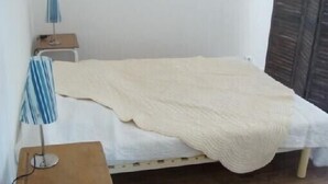 Une petite chambre avec un lit simple, une couette blanche, une table de chevet en bois avec une lampe et une porte en bois.
