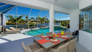 Un espace repas aménagé au bord de la piscine, avec une table dressée pour deux, offrant une vue sur la piscine, les palmiers et un bâtiment situé au loin.