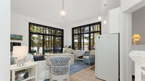 A modern kitchen with large windows, a dining area with wicker furniture, and a white refrigerator.