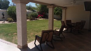 Un patio cubierto con una mesa, sillas, ventilador de techo y vistas a un jardín.