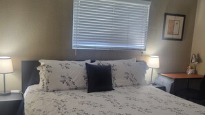 Un dormitorio con una cama, dos mesitas de noche, una lámpara de noche, un ventilador de techo, una ventana con persianas y un cuadro enmarcado en la pared.