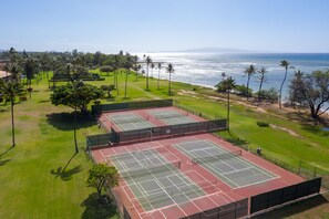 Vista aérea de un complejo de canchas de tenis con múltiples canchas, rodeado de vegetación y palmeras, con vista a una playa y el océano.