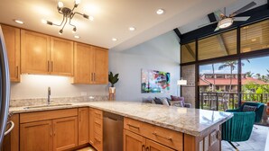 A modern kitchen with wooden cabinets, a granite countertop, and a dishwasher. There is a dining area with a view of palm trees and a building outside.