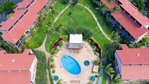 An aerial view of a hotel with a central swimming pool, surrounded by palm trees and a well-maintained lawn.