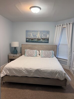 Een slaapkamer met een houten bed, een nachtkastje met een lamp, een schilderij van zeilboten en een raam met gordijnen.