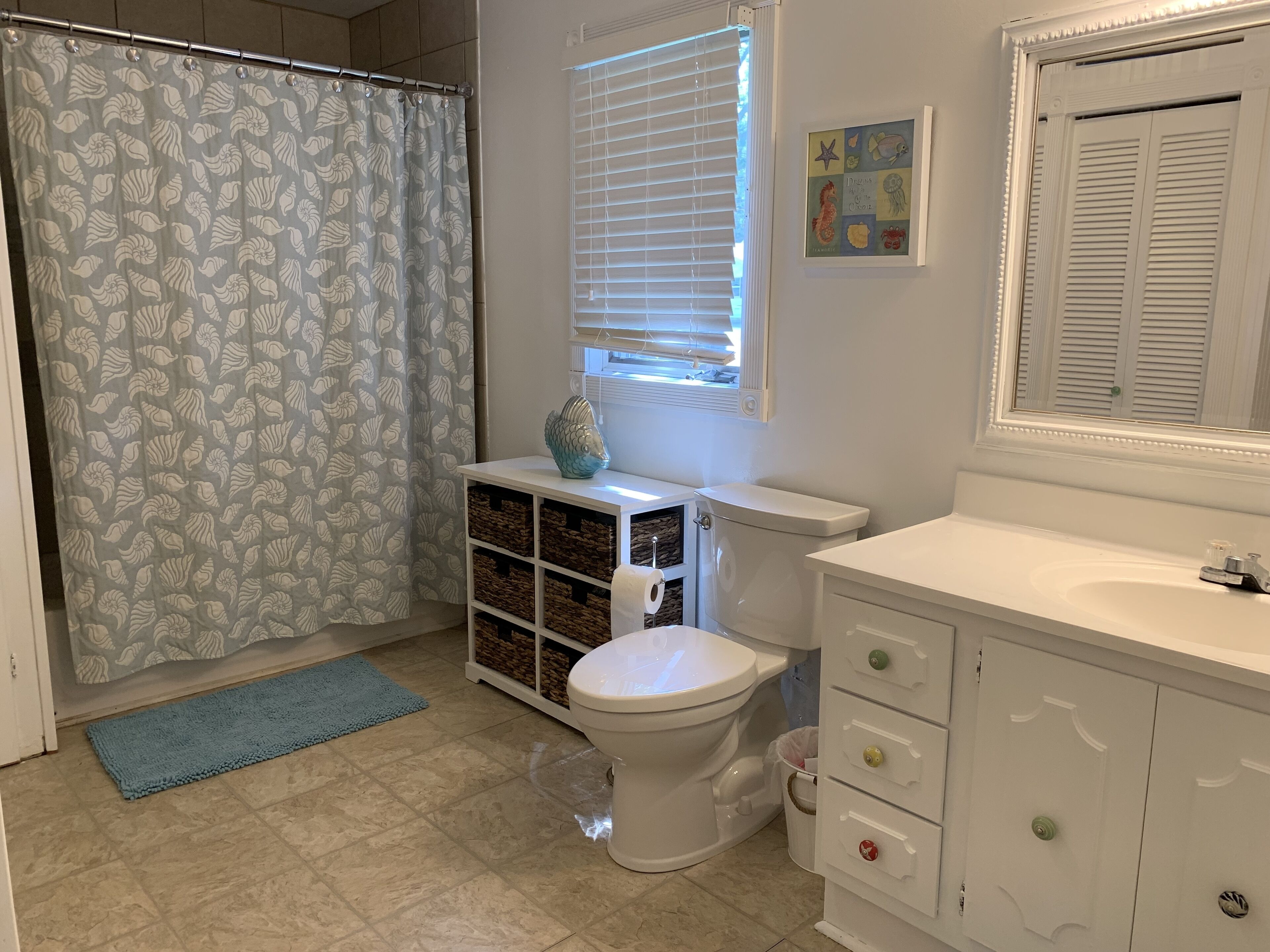 A bathroom with a toilet, a sink, a window with blinds, a shower curtain, and a decorative fish on the sink.