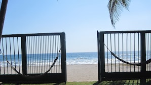 Blick durch ein Metallgatter auf einen Strand mit Wellen, wolkenlosem Himmel und einer Palme.