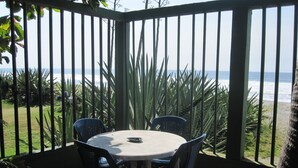 Ein überdachter Außenbereich mit Tisch und Stühlen mit Blick auf einen Strand, Palmen und das Meer.