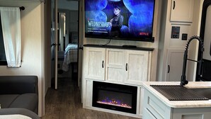Een compacte leefruimte met een flatscreen televisie boven een open haard, houten vloeren en een kitchenette.