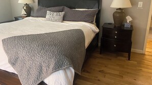 Un lit bien fait, avec une couverture et des oreillers gris, deux tables de chevet avec des lampes, une horloge murale et un panneau au-dessus du lit.