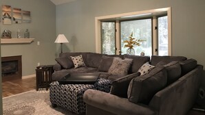 Un salon comprenant un canapé d’angle, un pouf à motifs, une cheminée et une grande fenêtre avec des stores.
