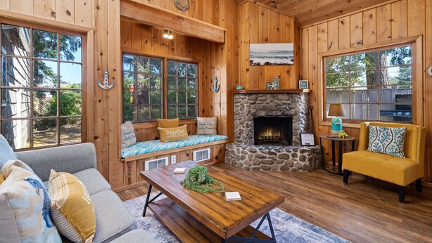 A cozy cabin interior with a stone fireplace, wooden walls, a large window, and a seating area with a sofa and a yellow armchair. - BP:~Walk2Beach/Cafe/Golf/Store&Pub~Hottub~Bikes~Firepit~PingPong~Fenced~Dog (Seaside)