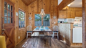 A wooden cabin interior with a dining table set for four, two chairs, a chandelier, and wooden cabinets. - BP:~Walk2Beach/Cafe/Golf/Store&Pub~Hottub~Bikes~Firepit~PingPong~Fenced~Dog (Seaside)