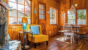 A cozy cabin interior with wooden walls, a stone fireplace, a yellow armchair, a dining table with chairs, and a chandelier. - BP:~Walk2Beach/Cafe/Golf/Store&Pub~Hottub~Bikes~Firepit~PingPong~Fenced~Dog (Seaside)
