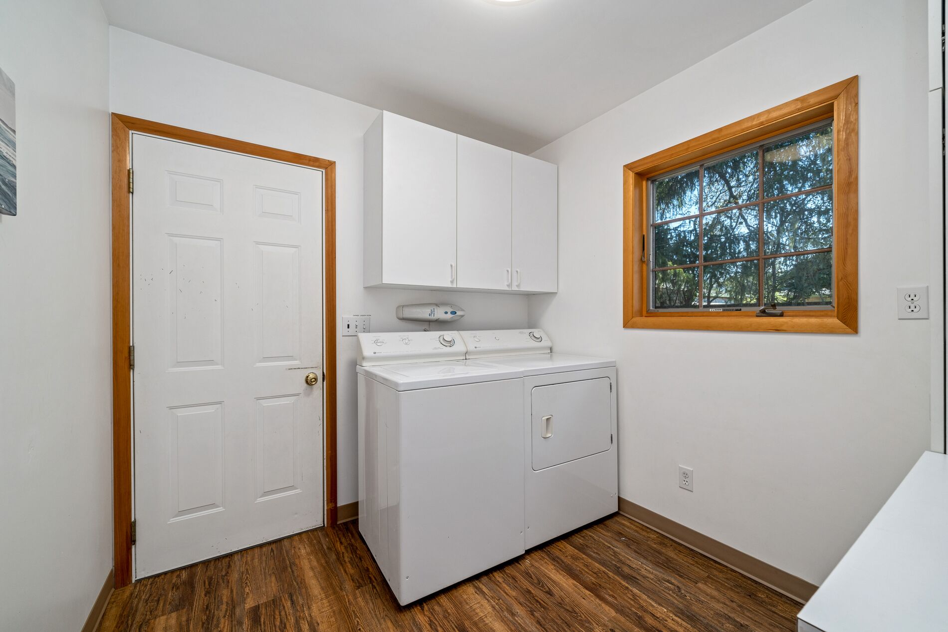 A laundry room with a washer and dryer, wooden flooring, white walls, and a window with a view of trees.