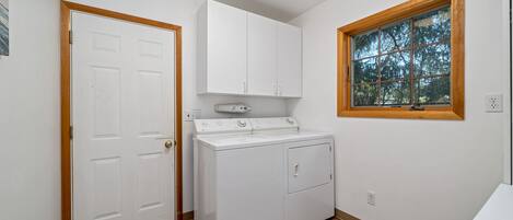A laundry room with a washer and dryer, wooden flooring, white walls, and a window with a view of trees.