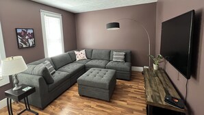 A living room with a sectional sofa, a small table, a lamp, a TV, and a wooden floor. - Cozy updated 3 BR home steps away from downtown, space to relax and enjoy.  (Holland)