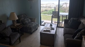 A living room with a sofa, armchair, and a coffee table. There is a lamp on a side table, and a model ship on the wall. The room has a large window with a view of the beach. - Direct Oceanfront Condo and Drive on the beach access!! (Saint Augustine)