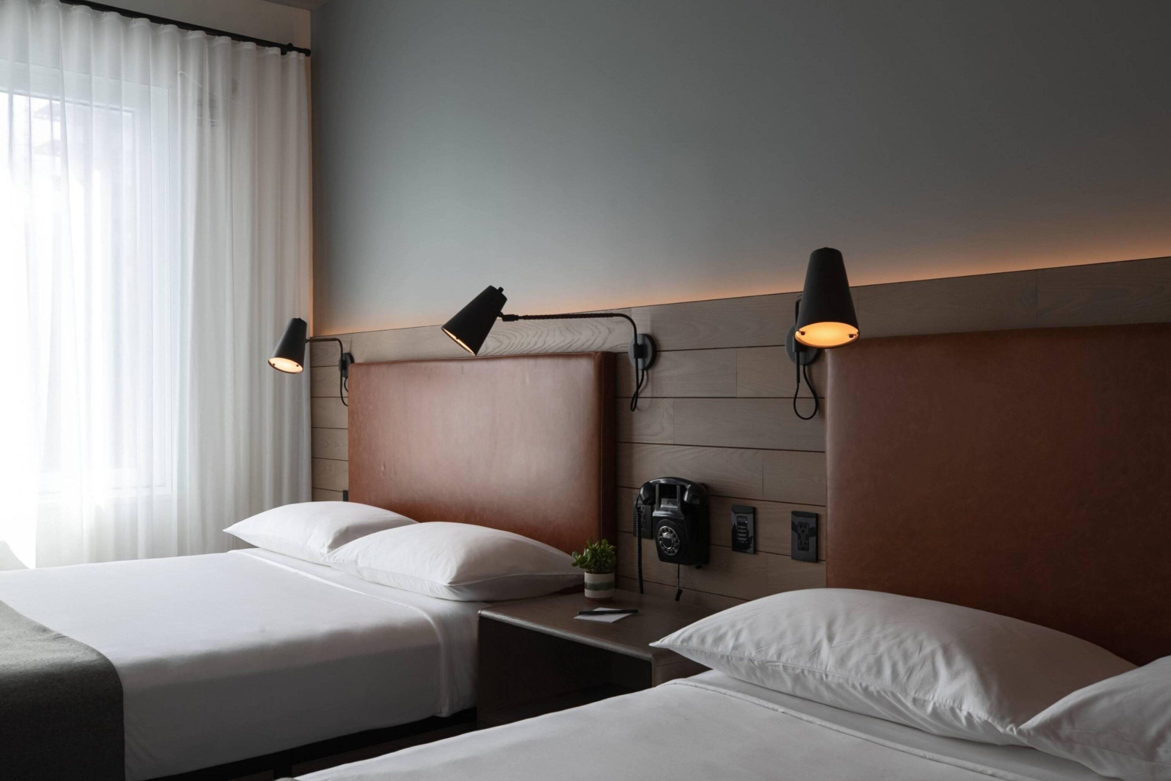 Quarto de hotel com duas camas, cabeceira de madeira, luminárias laterais, telefone e janela com cortinas.