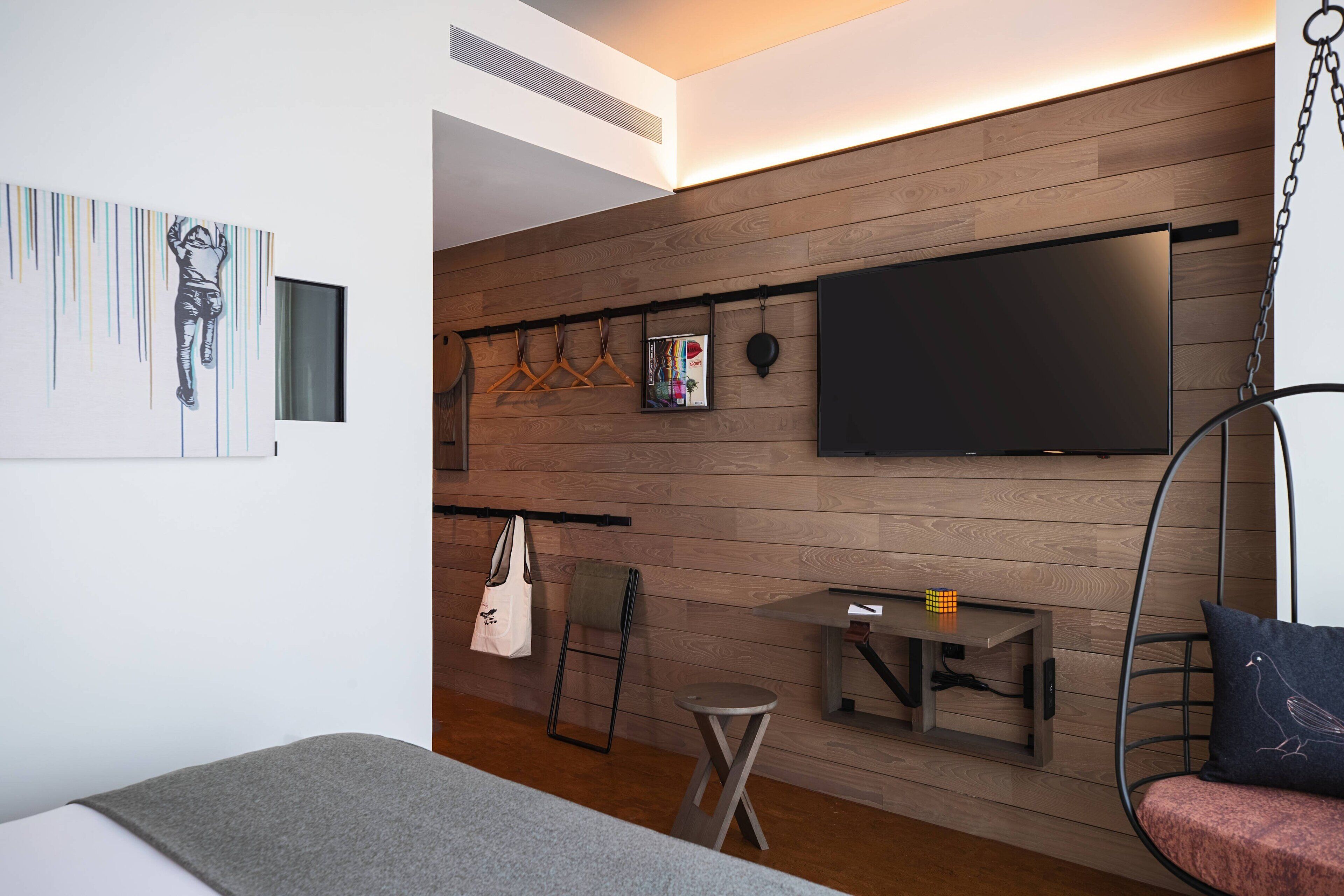 A modern room with a wooden wall, a flat-screen TV, a hanging chair, a small table with a puzzle, and a bed with a grey blanket.