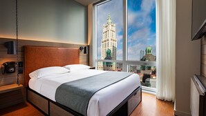 A hotel room with a large window, a bed, a wooden headboard, a bedside table with a telephone, and a view of a cityscape. - Moxy NYC Downtown (New York)