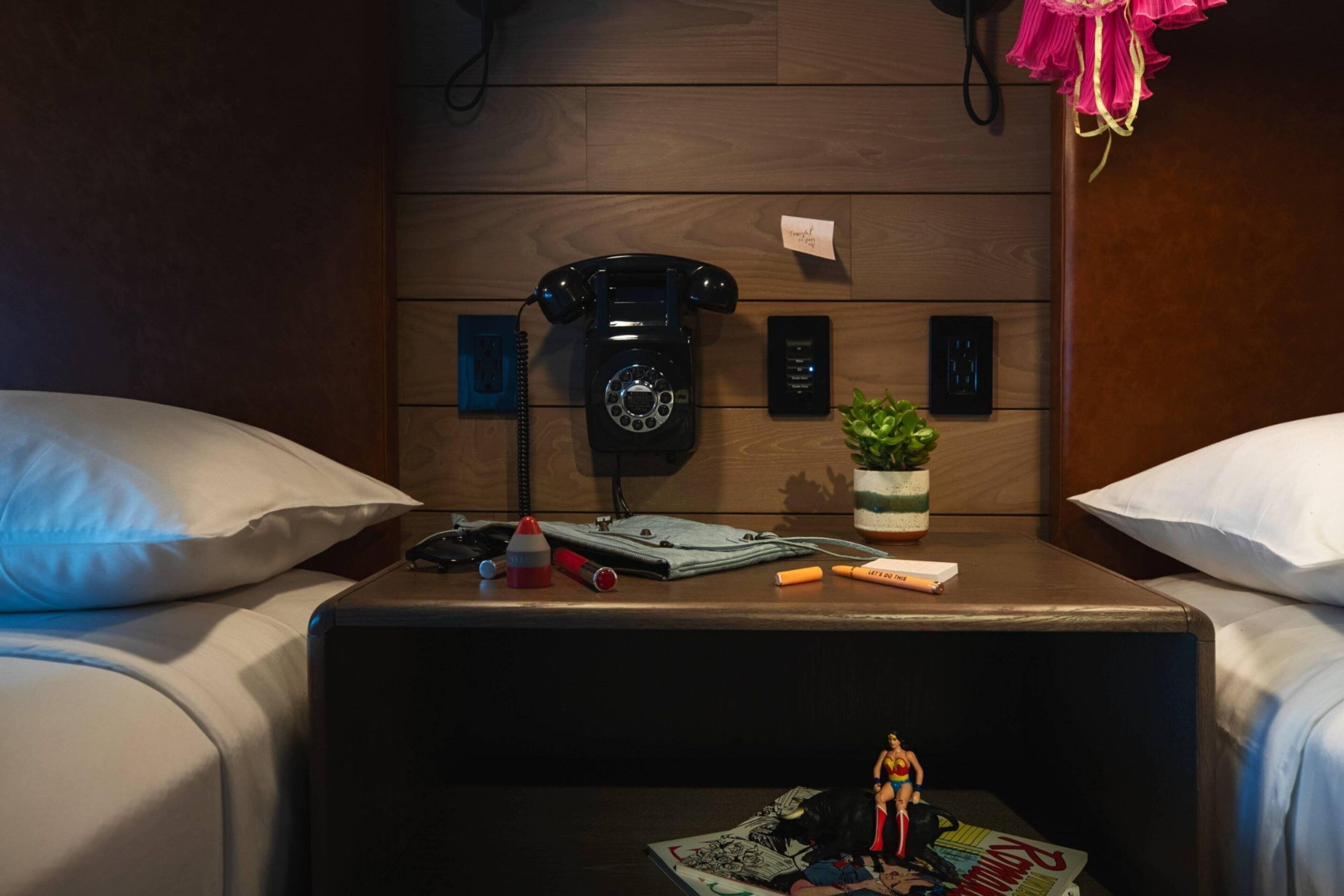 A hotel room with a wooden headboard, a black rotary phone, a small potted plant, and a magazine on the nightstand.