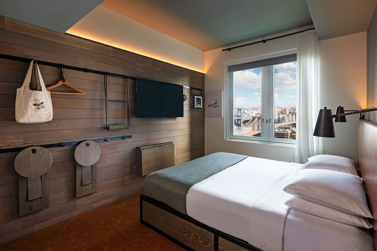 A modern hotel room with a bed, a TV, a view of the city, and a wooden wall design.