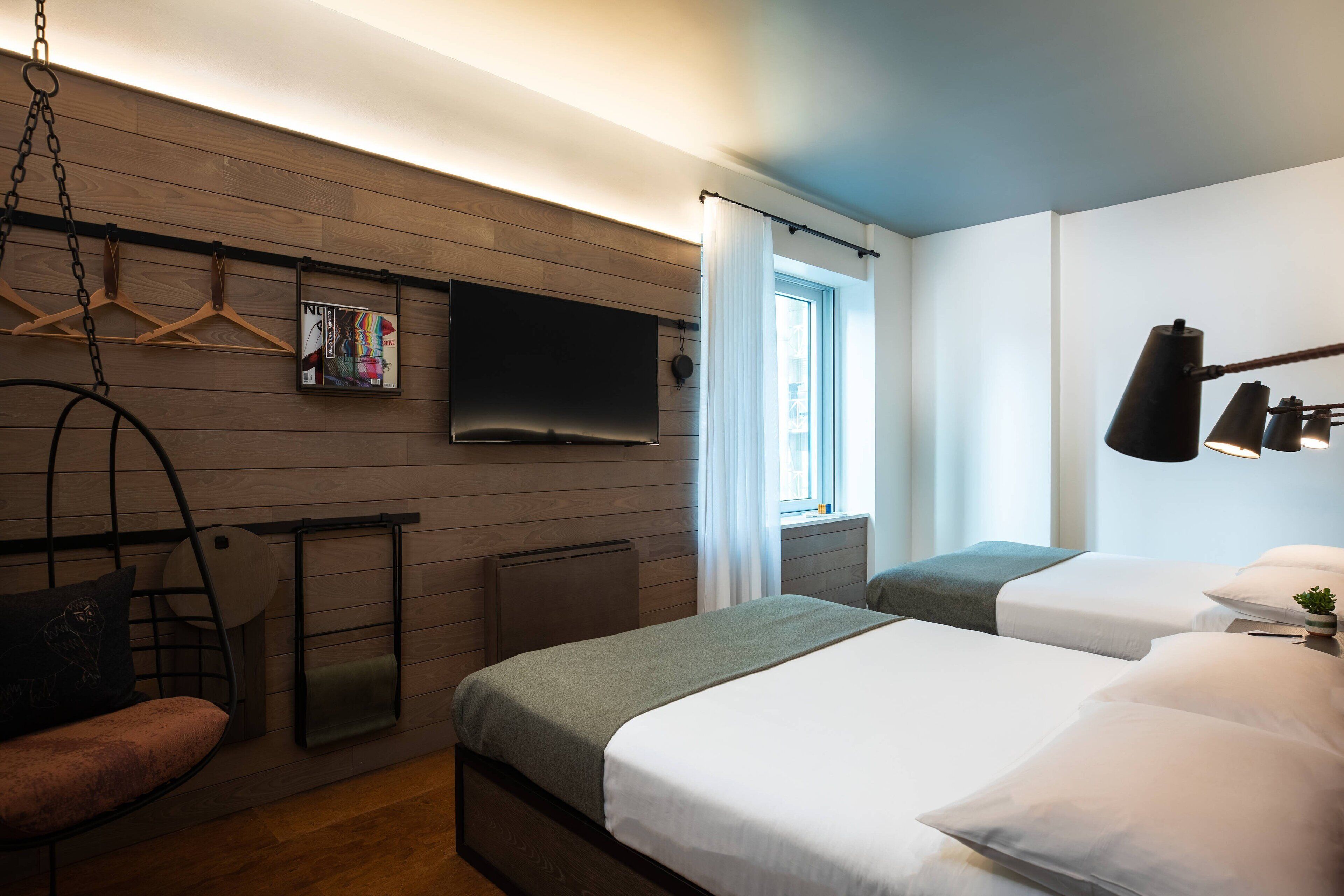 A hotel room with two beds, a TV mounted on the wall, a hanging chair, and a window with curtains.