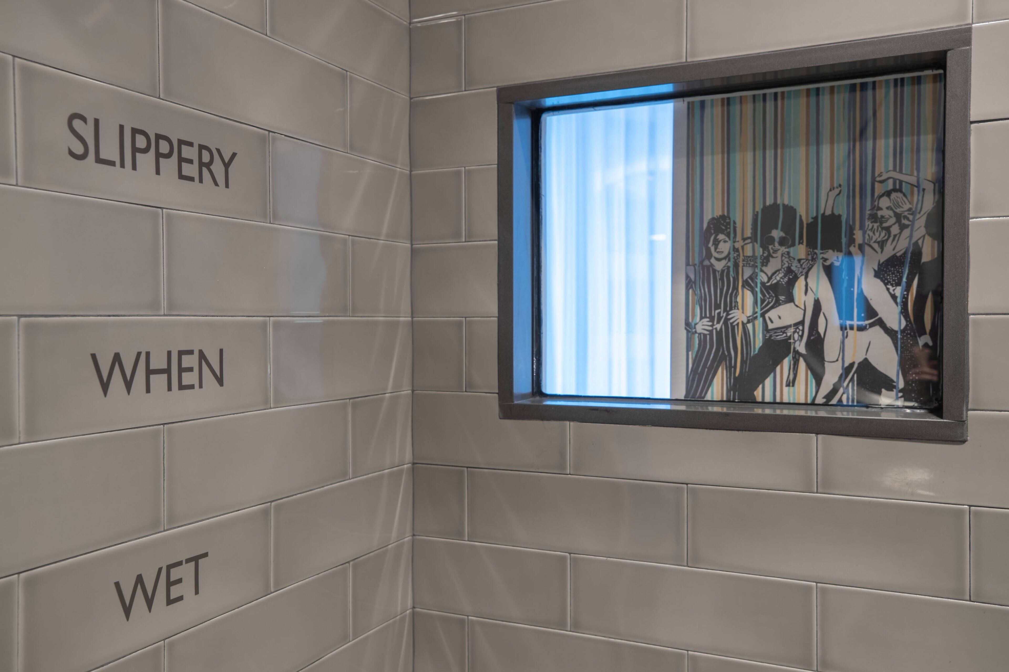 A tiled wall with the words "SLIPPERY WHEN WET" and a window with a striped pattern.