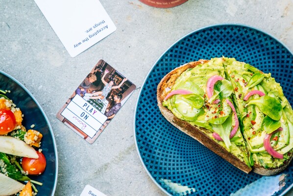 Una mesa con un plato azul que contiene un sándwich cubierto de aguacate, lechuga y cebollas rojas. También se ven cartas y un pequeño bol con ensalada.