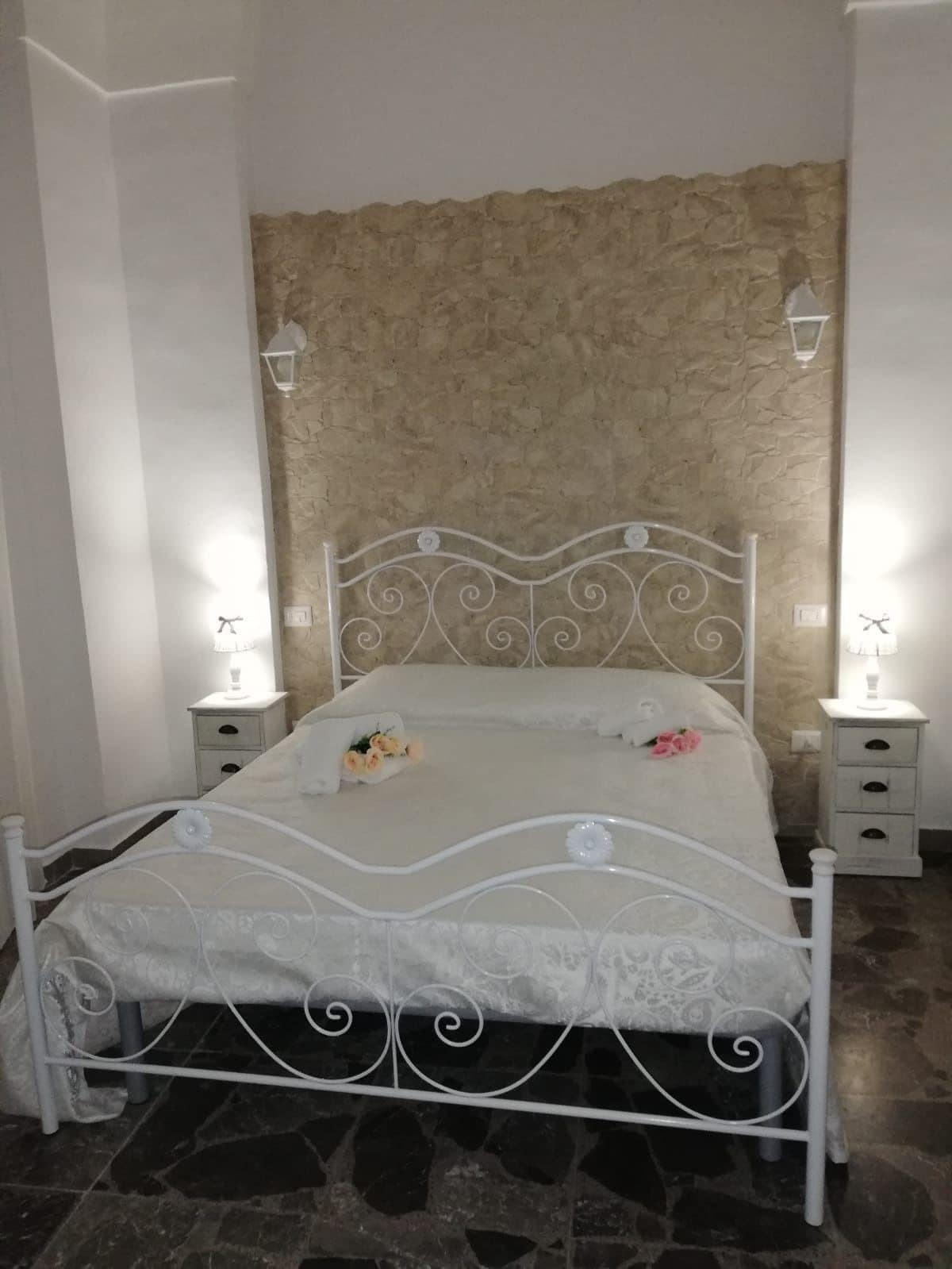 Ein Bett mit weißem Metallgestell vor einer Steinwand. Zwei Nachttische mit Lampen stehen daneben.